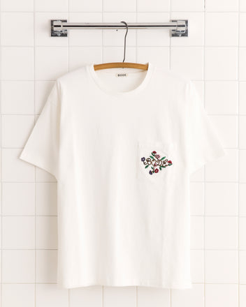 The Posy Pocket Tee—a white t-shirt featuring a flower pattern and floral pocket embroidery—hangs on a wooden hanger against a tiled wall.