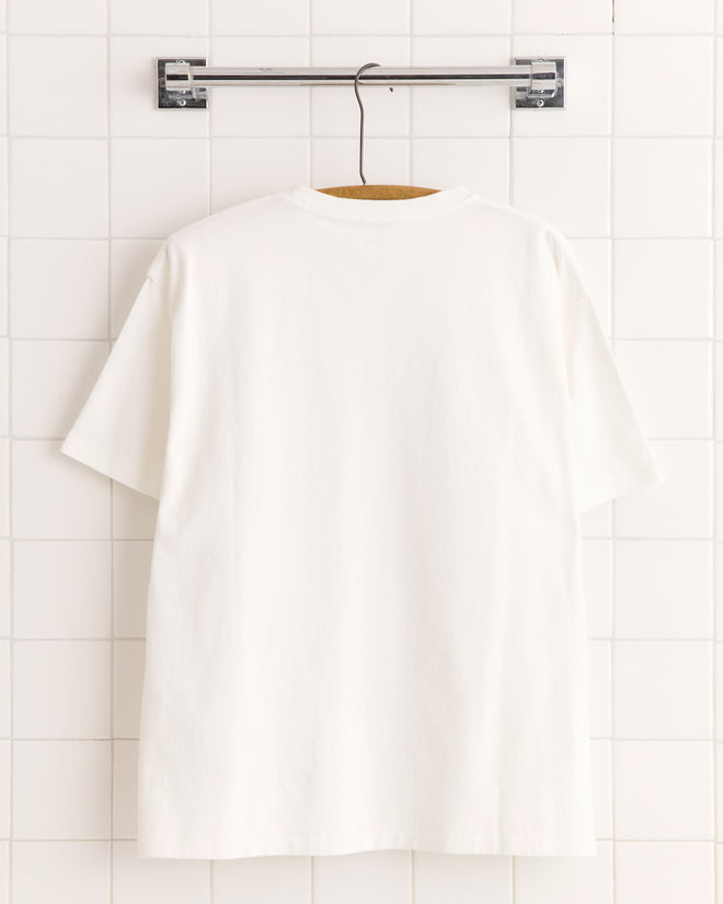 The Posy Pocket Tee hangs on a wooden hanger against a white tiled wall.