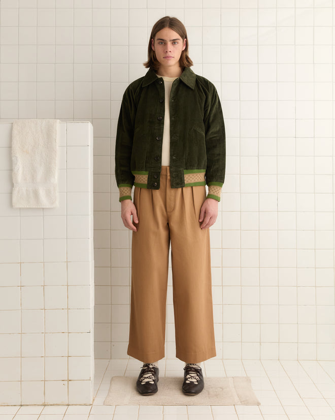 A person in a forest green Peerie Rib Banbury Jacket and brown pants stands in a tiled room, with a towel hanging on a partition.