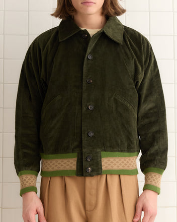 A person in a forest green Peerie Rib Banbury Jacket and tan pants stands against a tiled wall.