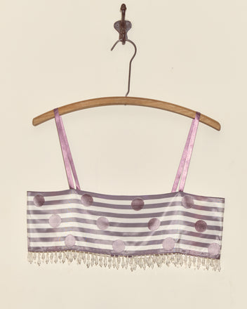 The Purple Note Bra - S, featuring purple polka dots and stripes with beaded fringe, crafted from Staron jacquard ribbons sourced from a French couture supplier, hangs on a wooden hanger against a plain wall.