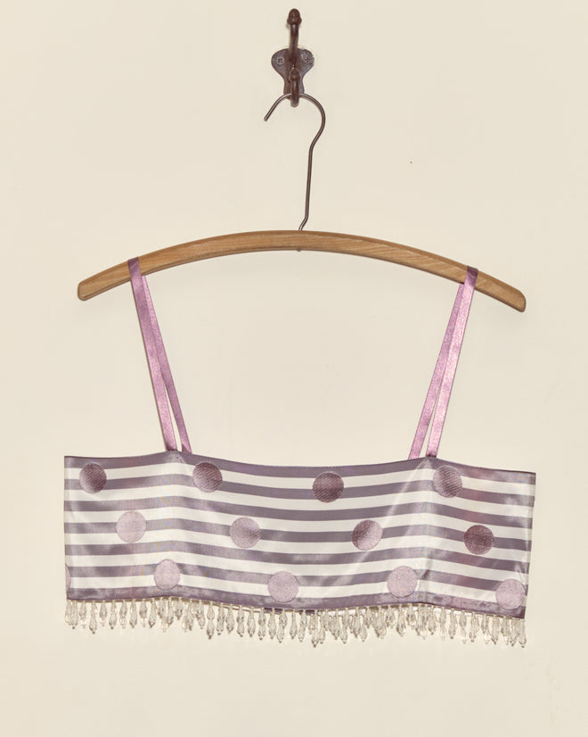 The Purple Note Bra - S, featuring purple polka dots and stripes with beaded fringe, crafted from Staron jacquard ribbons sourced from a French couture supplier, hangs on a wooden hanger against a plain wall.