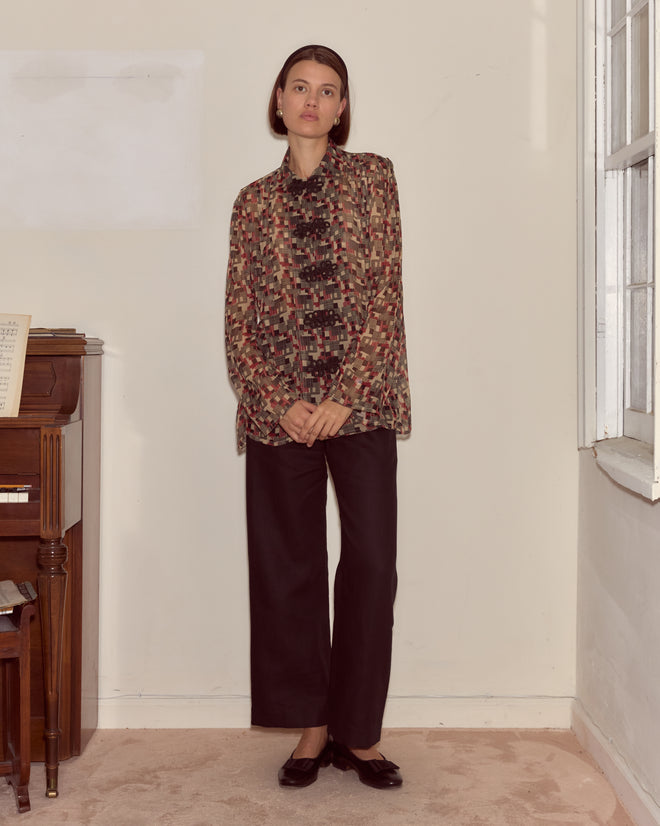 A woman in the Quadrille Blouse, featuring a 1930s-inspired geometric print, and black pants stands indoors by a piano and window.