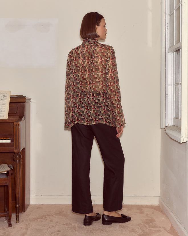 Woman standing indoors, wearing the Quadrille Blouse with a 1930s-inspired geometric print, paired with black pants and loafers, facing away from the camera.
