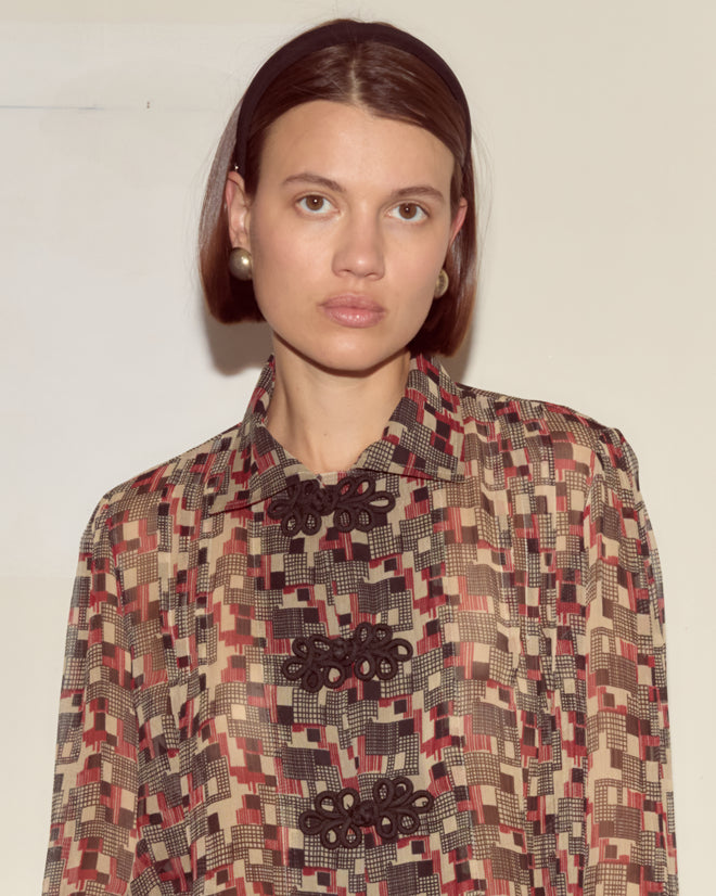 A woman with straight brown hair wears the Quadrille Blouse, featuring a 1930s-inspired geometric print, and stands against a beige wall looking at the camera.
