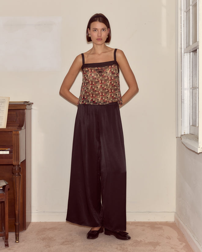 A woman stands indoors by a piano and window, wearing the Quadrille Tank with a geometric pattern and wide-leg black pants.