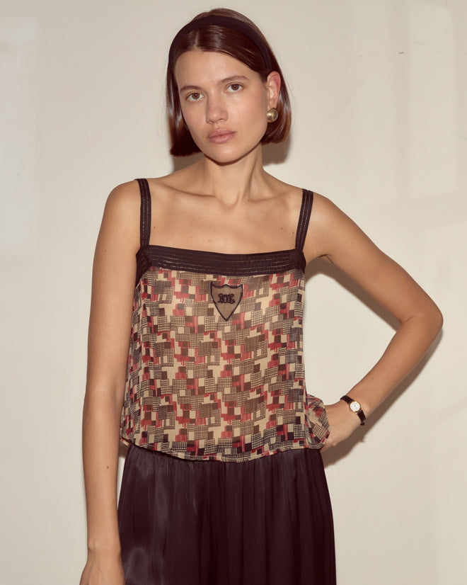 A woman wears the Quadrille Tank, featuring a geometric pattern, paired with a black skirt and stands against a plain light-colored wall.