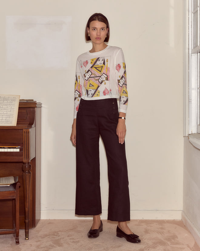 A woman with short brown hair stands indoors by a piano, wearing the Queen Of Hearts Long Sleeve Tee made from 100% cotton and black pants.