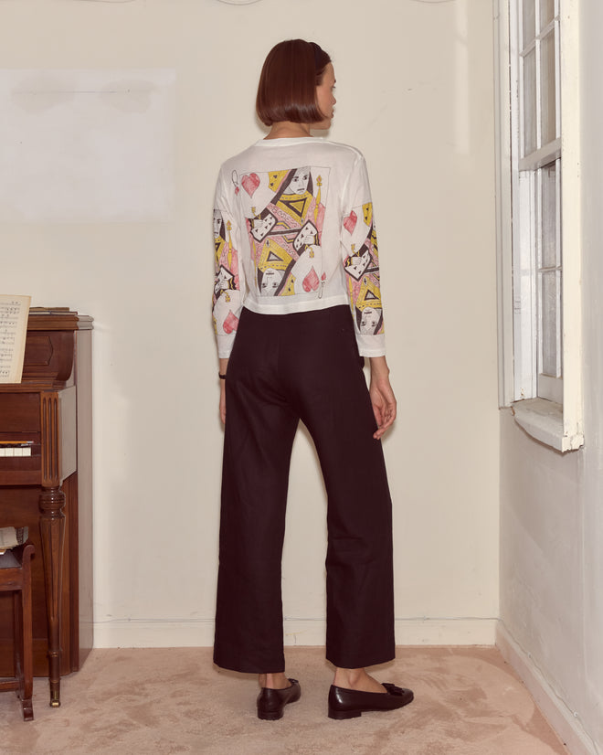 A woman stands indoors near a piano and window, wearing the Queen Of Hearts Long Sleeve Tee (100% cotton) and black pants, evoking an Alice in Wonderland vibe.