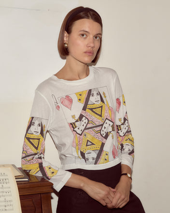 A woman wearing the Queen Of Hearts Long Sleeve Tee, featuring playing card queen designs, stands beside a piano with sheet music.