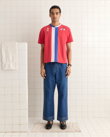 A person in a Racing Stripe Tee and jeans stands on a bath mat in a tiled room, with a towel draped over the partition.