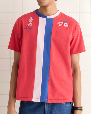 A person confidently stands in front of a tiled wall, wearing the Racing Stripe Tee featuring vertical stripes and embroidered figures, inspired by the 1980s.