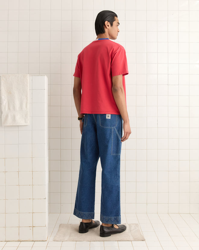 In a tiled room with a towel on the wall, someone in blue jeans and the Racing Stripe Tee red shirt stands facing away.