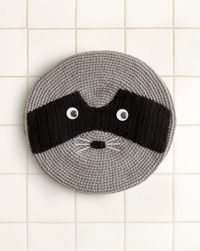 Raccoon Beret on a crocheted face rug with a black mask and googly eyes, set against a tiled background.