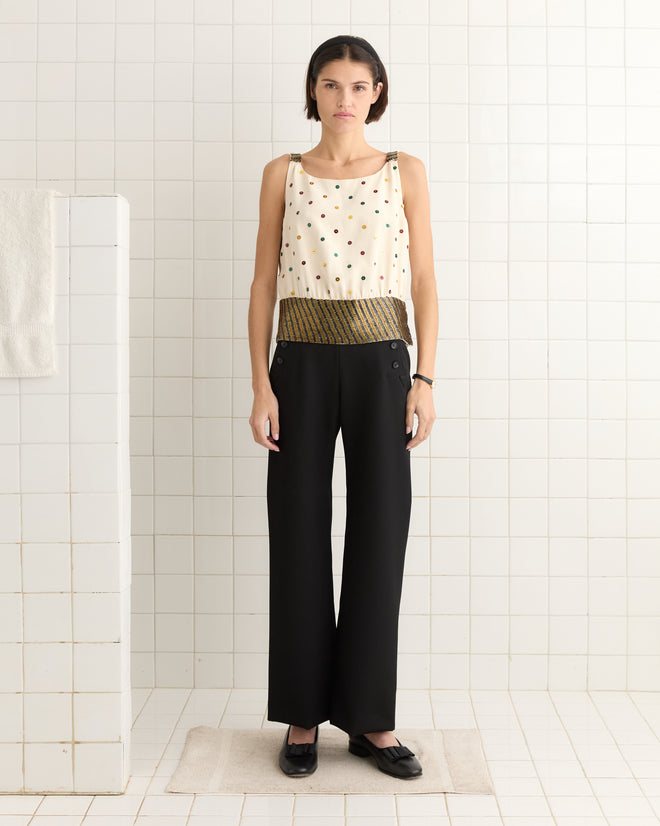 A woman in a Raffle Top and black pants stands gracefully on a rug, surrounded by the pristine elegance of a white-tiled room.