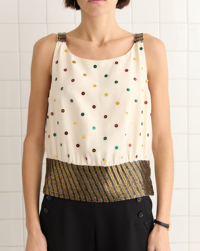 A person in a sleeveless Raffle Top, featuring multicolored dots, a gold-striped hem, and shimmering sequins, stands against a tiled wall.