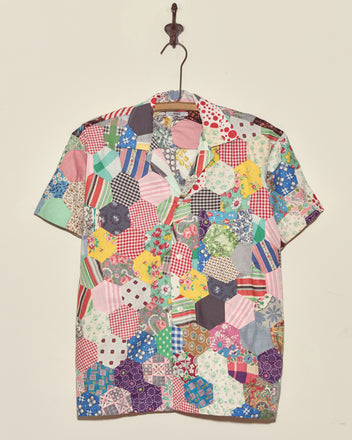 The Rainbow Fish Quilt Shirt - XS/S, a short-sleeved top featuring colorful, assorted vintage fabric patterns, hangs on a wall-mounted hook.