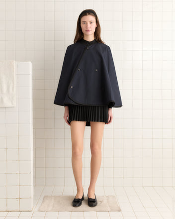 A woman in a navy rain cape and skirt stands on a tiled floor, facing forward. The Waxed Herringbone Rain Cape, made from water-resistant waxed cotton, adds an air of sophistication to the scene.