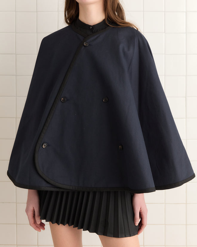 Against a tiled wall, an individual donning the Navy Waxed Herringbone Rain Cape and pleated skirt showcases both style and protection with its water-resistant waxed cotton.