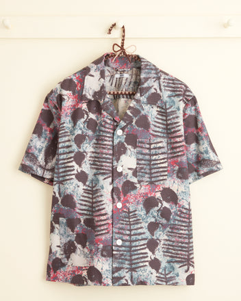 The Rainforest Shirt - XS/S, adorned with abstract black, blue, and pink botanical patterns and styled with short sleeves and buttons, hangs on a hook against a cream-colored wall backdrop.