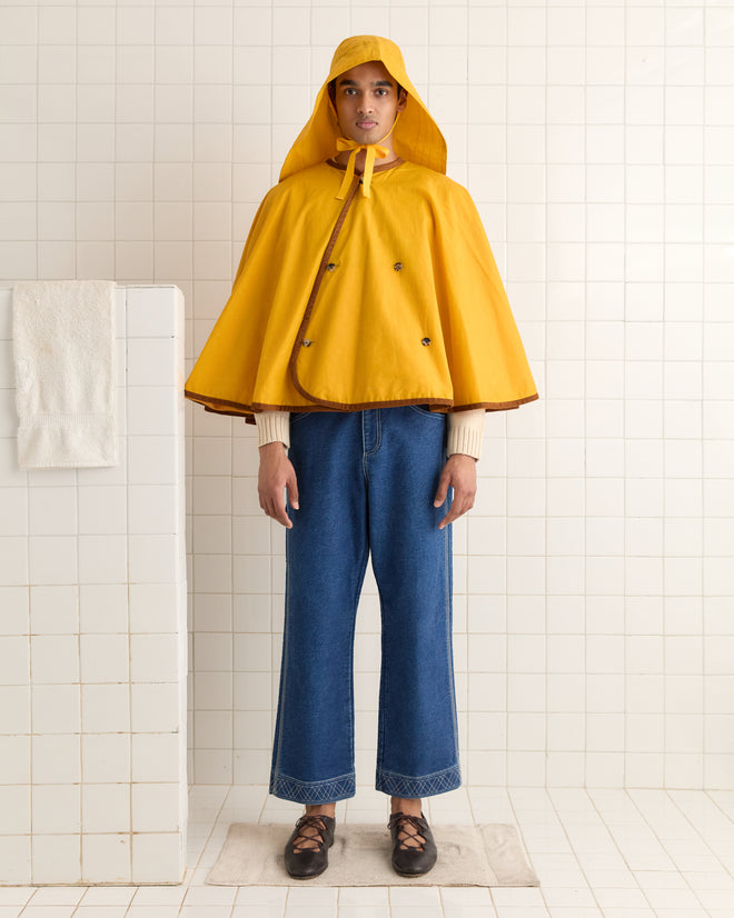 In a white tiled room, someone in jeans and black shoes pairs their yellow rain poncho with a Waxed Rain Hat - Yellow for the perfect blend of practicality and style.