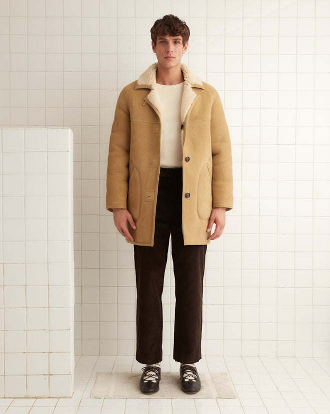 A person in a beige Ranch Coat, black pants, and white shoes stands against a white tiled wall.