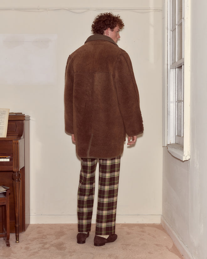 A person wearing the Ranch Coat - Brown and plaid pants stands facing a wall near a piano and window in a carpeted room.