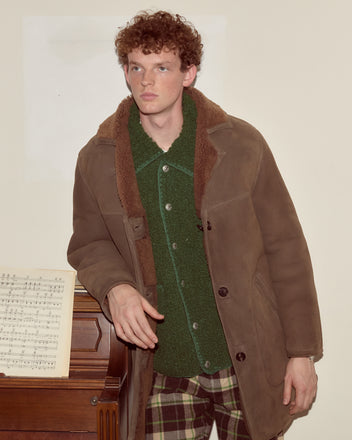 A man in a brown Ranch Coat stands by a piano with sheet music, wearing plaid pants and looking away.