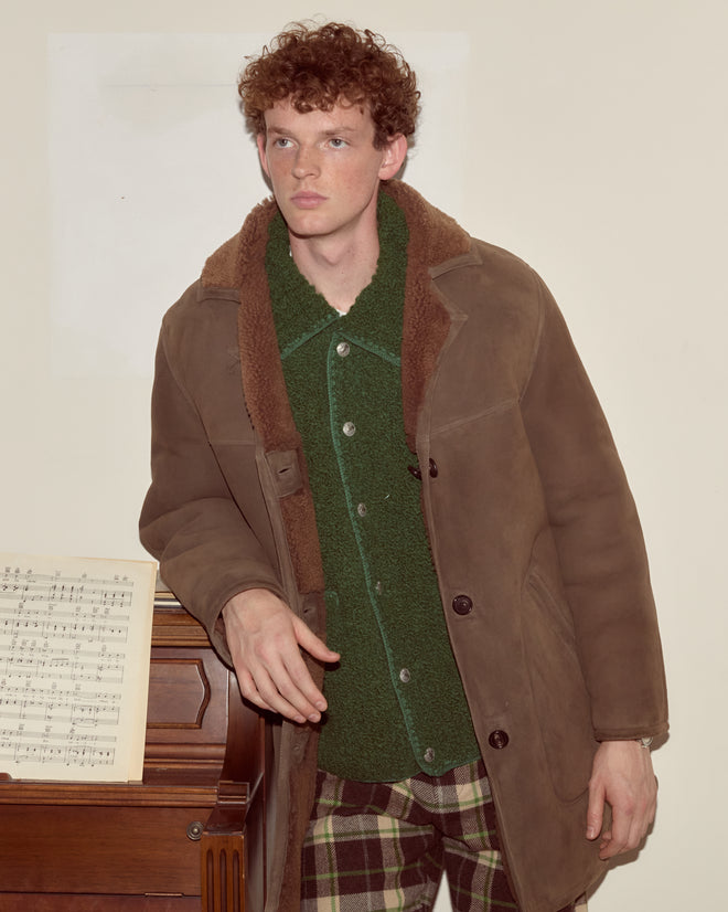 A man in a brown Ranch Coat stands by a piano with sheet music, wearing plaid pants and looking away.