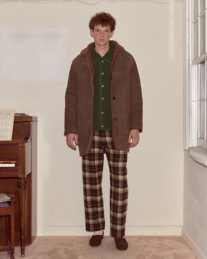 A man stands indoors near a piano wearing a green shirt, plaid pants, brown shoes, and the Ranch Coat - Brown.