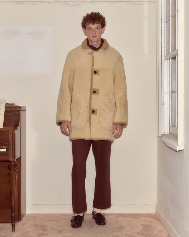 A person wearing the Ranch Coat and brown pants stands indoors beside a piano and a window.