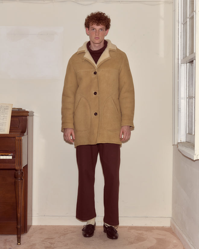 A man wearing a Ranch Coat and brown pants stands indoors next to a piano and a window.