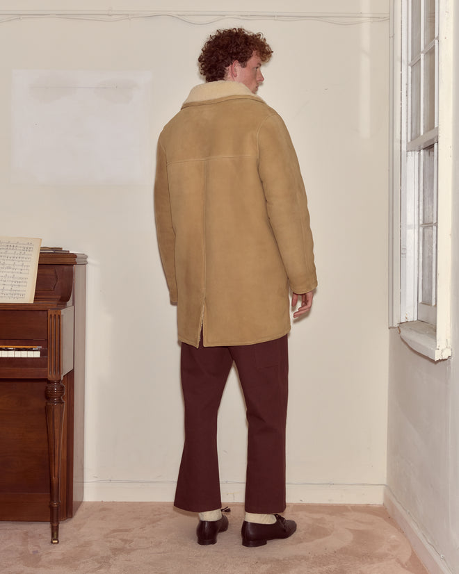 A man in a tan Ranch Coat and brown pants stands facing away in a room with a piano and window.