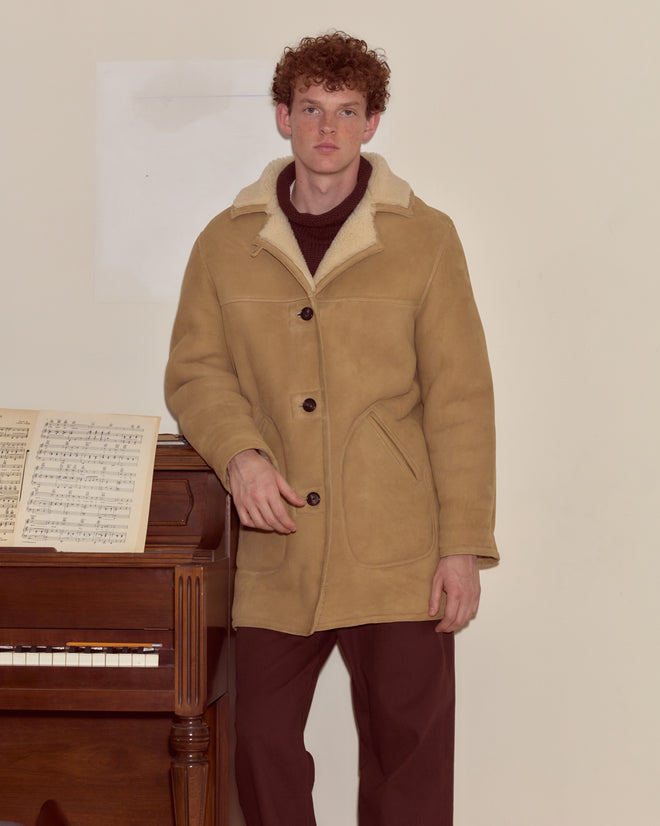 A man wearing the Ranch Coat stands beside a piano with sheet music, set against a plain wall.