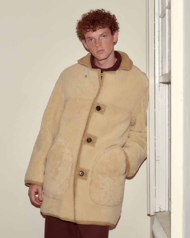 A person with curly red hair stands indoors by a window wearing the Ranch Coat, a 1950s reproduction shearling jacket.