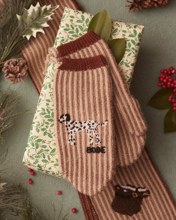 Ranch Mittens, Fair Trade Certified, rest atop a wrapped gift adorned with holly, pinecones, and berries.