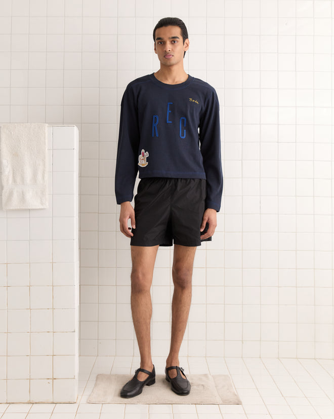A person in a navy Rec Pullover and black shorts stands in a tiled room with a towel hanging nearby.