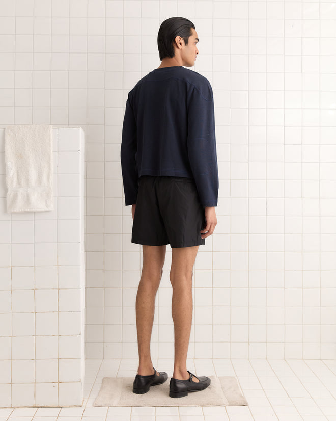 A person in a Rec Pullover - Navy and shorts stands in a tiled room, facing a wall with a towel hanging.