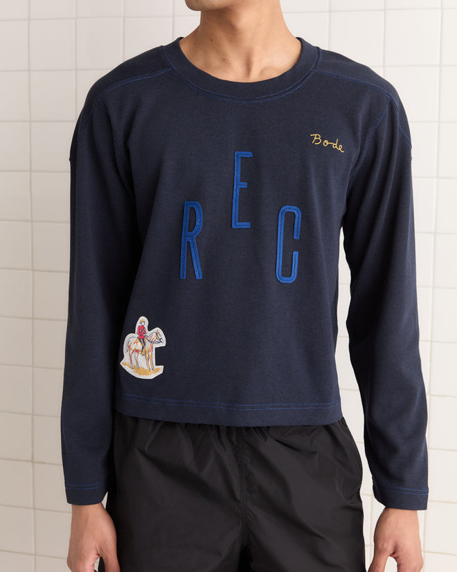 Someone wears a Rec Pullover in navy, featuring "REC" and an embroidered figure, made of heavyweight fabric, paired with black shorts.