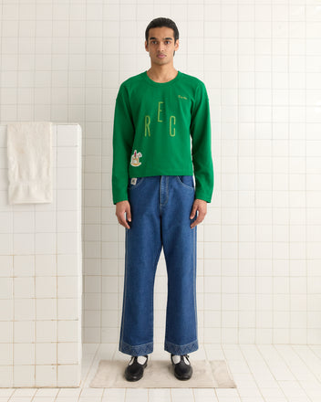 Wearing a Rec Pullover in green and blue jeans, an individual stands in a white-tiled room, where a basketball jersey is draped casually over the towel rack nearby.