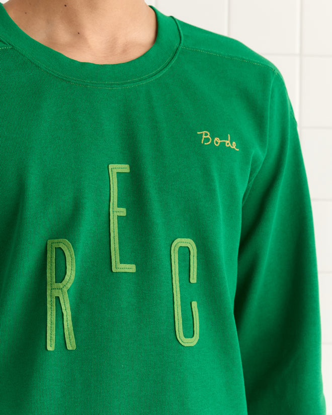 A person is wearing the Rec Pullover - Green, featuring "REC" in bold letters and a smaller embroidered name on the chest, reminiscent of classic vintage jerseys.