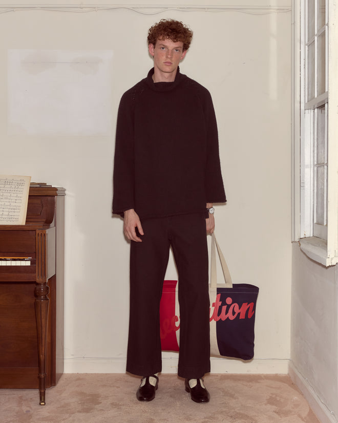 A person in loose black clothing stands indoors by a piano, holding a Recreation Tote and an American Bicentennial tote near an open window.