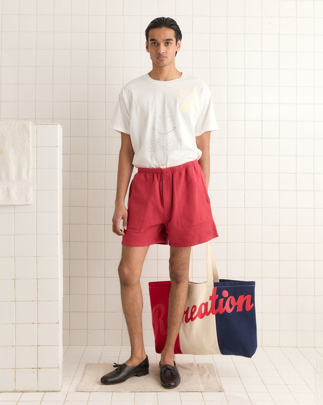 In a tiled room, a person in a white shirt and red shorts stands confidently, clutching the stylish Recreation Tote.