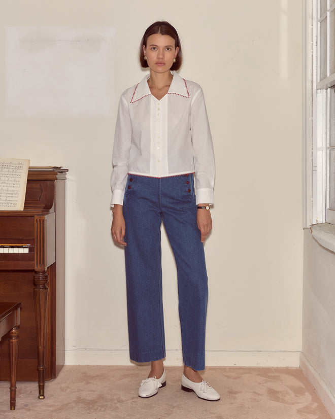 A woman stands indoors in the Redbud Top, blue pants, and white shoes next to a piano and a window.