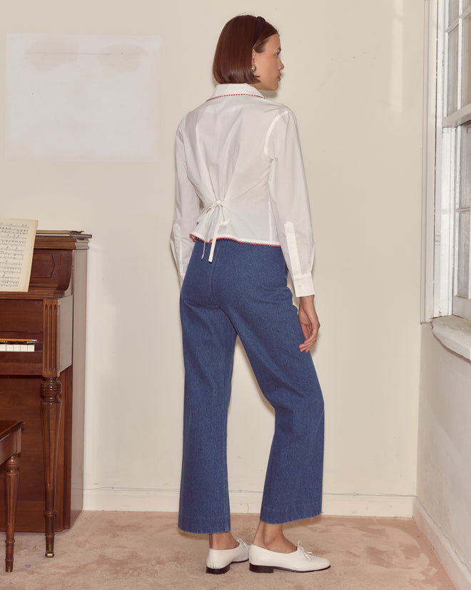 A woman in a Redbud Top and blue jeans stands by a window, facing away from the camera, near a piano.