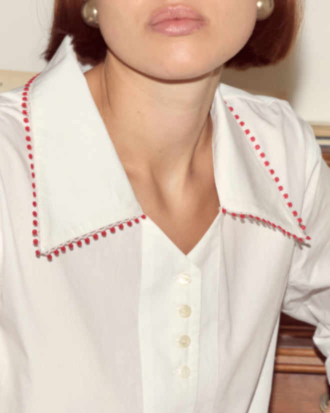 A woman is shown from the shoulders up wearing the Redbud Top, a white cotton poplin blouse with red lace trim, paired with pearl earrings.