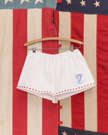 Red Star Tap Shorts hang on a wooden hanger with an American flag backdrop—white shorts with red star trim and a blue portrait, ideal for Liberty costumes or your Fourth of July outfit.