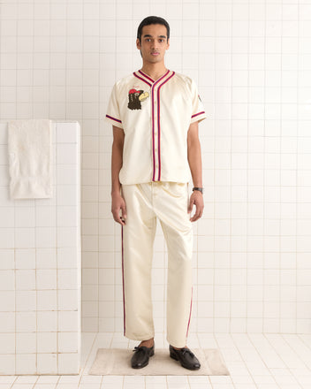 In a tiled room, a person wears the Redtail Baseball Shirt, featuring a cream color with red trim that channels 1960s style. A towel hangs neatly on a rack nearby, enhancing the vintage ambiance.