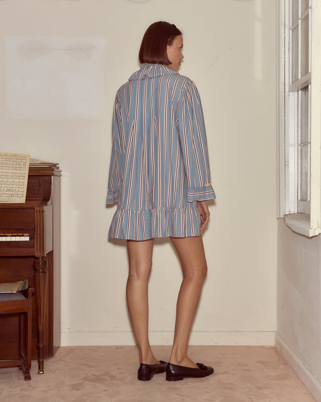 A woman wearing the Regatta Stripe Dress stands near a piano, facing away, in a softly lit room with a window.