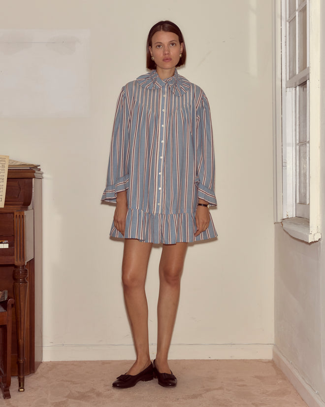 A woman wearing the Regatta Stripe Dress stands indoors by a piano and window, gazing at the camera.